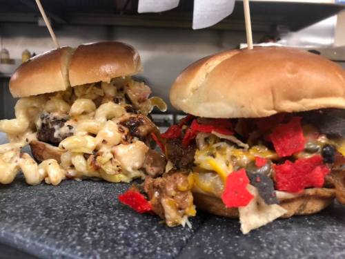 Two gourmet sandwiches on a slate platter: one with mac and cheese, the other topped with chips and meat.