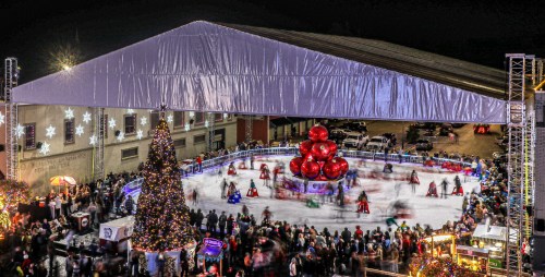 A festive outdoor ice skating rink under a large tent, surrounded by holiday decorations and a Christmas tree.