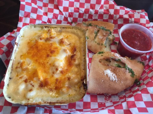 A tray with baked macaroni and cheese, stuffed breadsticks, and marinara sauce, all on a red and white checkered paper.