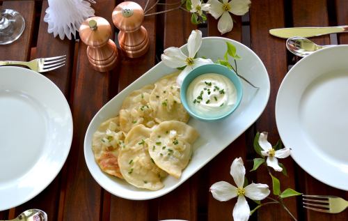 A plate of pierogi topped with chives, served with a small bowl of sour cream, surrounded by flowers on a wooden table.