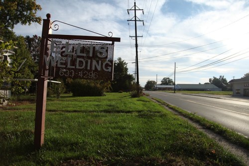 A metal sign for "Elli's Welding" stands by a grassy area next to a road, with power lines in the background.