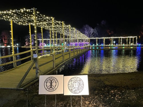 A lit wooden bridge over water at night, with festive lights and signs for a club and QR code in the foreground.