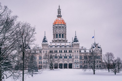 The Connecticut State Capitol building covered in snow, featuring a golden dome and intricate architectural details.