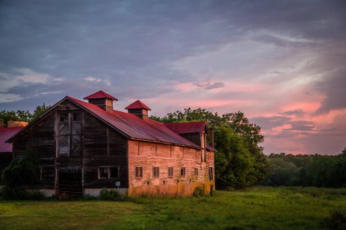 A rustic barn with a red roof stands in a grassy field under a colorful sunset sky.