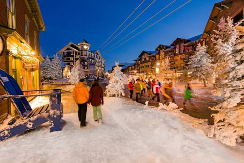 A snowy village scene at dusk, with people walking and colorful buildings illuminated by warm lights.