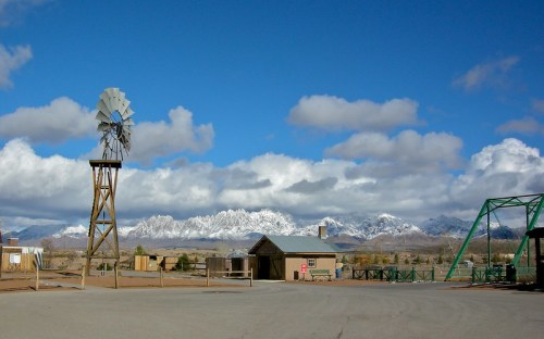 A windmill stands beside a building with snowy mountains and a blue sky in the background, featuring fluffy white clouds.