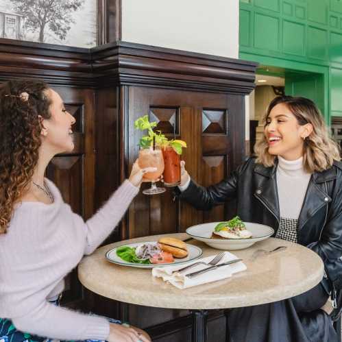 Two women sit at a table in a bright café, enjoying drinks and food, smiling and chatting.