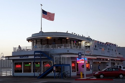 A seafood restaurant with an American flag, seagulls on the roof, and colorful neon signs at dusk.