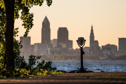 A scenic view of a city skyline at sunset, with a telescope in the foreground and trees framing the scene.