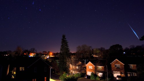 A night sky filled with stars, featuring a bright meteor streaking over a quiet suburban neighborhood.