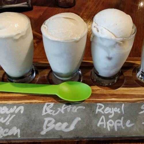 Four small glasses of ice cream in different flavors, displayed on a wooden tray with a green spoon.