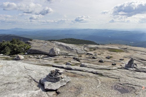 A rocky mountain landscape with scattered boulders and a panoramic view of rolling hills under a cloudy sky.