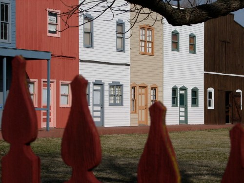 Colorful wooden buildings in a row, viewed through a red picket fence, with a grassy area in front.