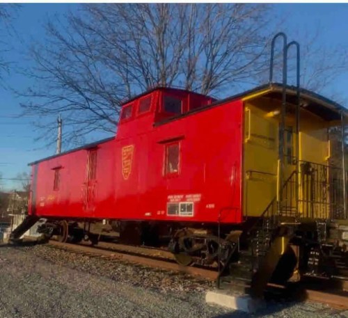 A bright red and yellow caboose on a railway track, surrounded by bare trees and a clear blue sky.