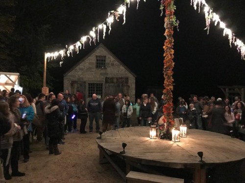 A crowd gathers around a decorated stage with lanterns, surrounded by trees and a rustic building at night.