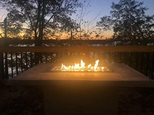 A fire pit with flames glows against a sunset backdrop by a lake, surrounded by trees.