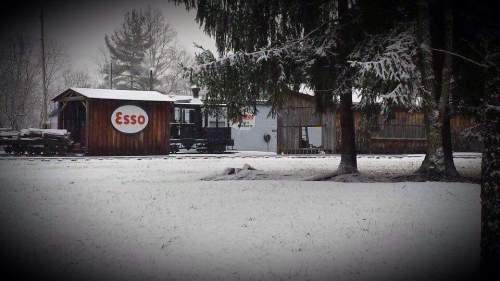 A snowy landscape featuring a rustic Esso gas station surrounded by trees.