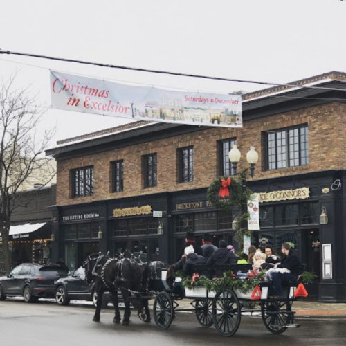 A horse-drawn carriage passes by a brick building with a festive banner reading "Christmas in Excelsior."