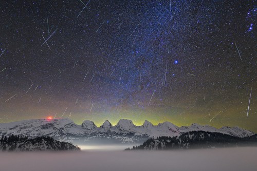 A starry night sky with shooting stars above snow-capped mountains and a foggy valley below.
