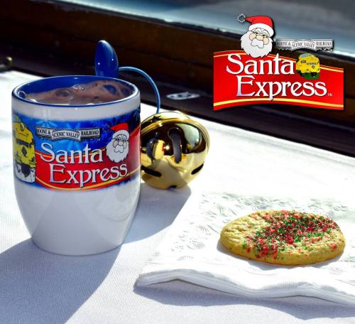 A festive mug of hot chocolate next to a decorated cookie on a napkin, with a jingle bell in the background.