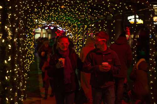 A couple walks under a canopy of colorful holiday lights, holding cups and wearing festive glasses.