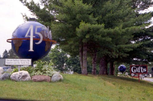 A large globe marker indicating the 45th parallel, with a nearby sign for gifts, surrounded by trees and grass.