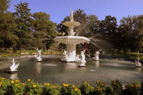 A white fountain with sculptures surrounded by greenery and flowers, set against a clear blue sky.