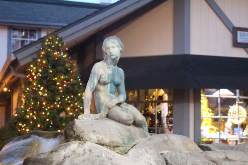 A weathered mermaid statue sits on rocks near a festive, decorated storefront with a Christmas tree.