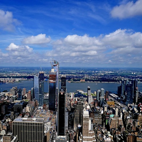 Aerial view of a city skyline with modern skyscrapers, under a blue sky with clouds, overlooking a river.