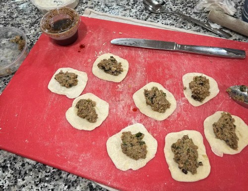 Dough circles on a red cutting board, each filled with a savory mixture, with a jar of sauce and a knife nearby.