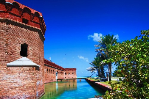 A historic brick fort beside a calm waterway, with palm trees and a clear blue sky in the background.