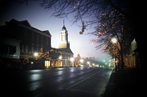 A quiet street at dusk, featuring a clock tower and buildings adorned with festive lights.