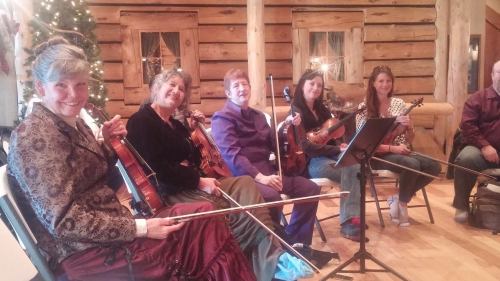 A group of six musicians seated indoors, playing violins, with a rustic wooden backdrop and festive decorations.