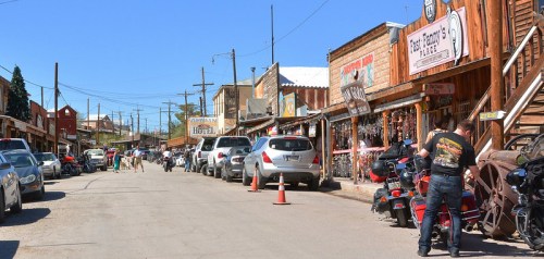 A historic street lined with wooden buildings, parked cars, and motorcycles under a clear blue sky.