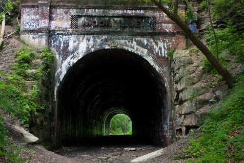 A dark, graffiti-covered tunnel surrounded by lush greenery and rocky walls, leading into a bright opening.