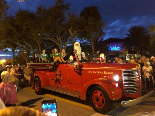 A vintage red fire truck in a parade, with people and Santa waving to the crowd under a twilight sky.