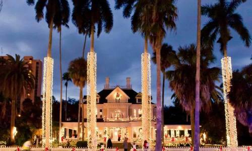 A grand mansion adorned with festive lights, surrounded by tall palm trees under a twilight sky.