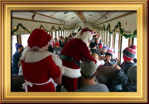 Two people dressed as Santa and Mrs. Claus interact with children on a decorated train filled with festive decorations.
