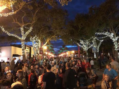 A bustling street scene at night, filled with people and decorated with festive lights on trees.