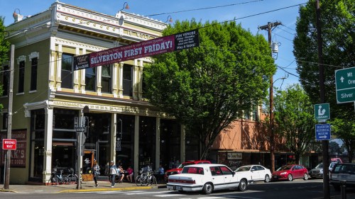 A vibrant street scene featuring a historic building with a banner for "Silverton First Friday" and green trees.