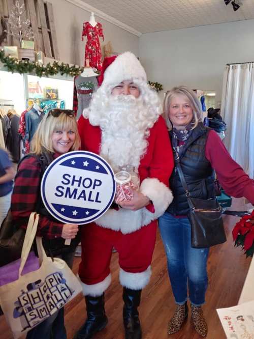 Two women pose with Santa Claus, holding a "Shop Small" sign in a festive store decorated for the holidays.