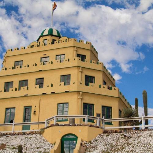 A yellow, multi-level building with a green dome and flag, surrounded by desert landscape and blue sky.