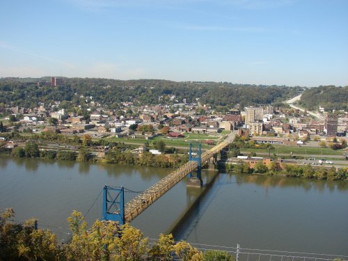 Aerial view of a bridge spanning a river, with a town and hills in the background under a clear blue sky.