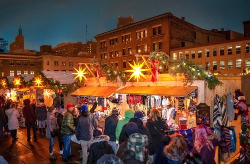 A bustling holiday market at night, with festive lights and people shopping at wooden stalls.