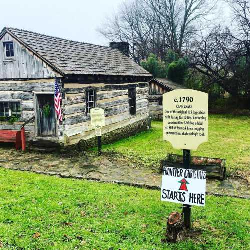 Historic log cabin from 1790 with a sign reading "Frontier Christmas Starts Here" in a grassy area.