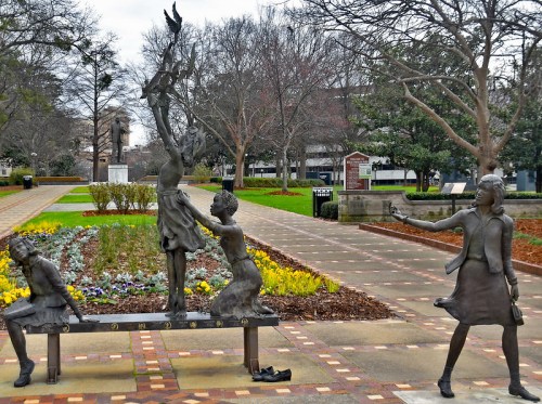 Three bronze sculptures of children playing in a park, surrounded by flowers and trees, with a pathway in the background.