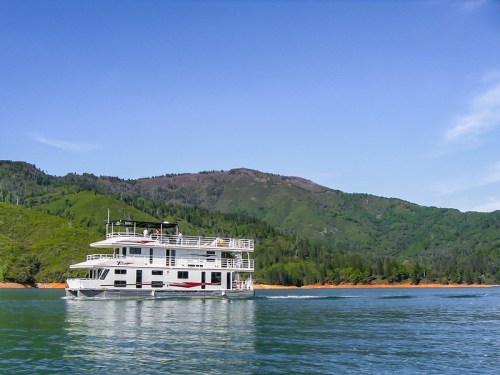 A houseboat glides across a calm lake, surrounded by lush green hills under a clear blue sky.