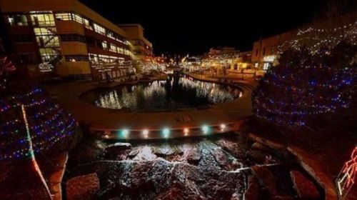 A nighttime view of a pond surrounded by colorful holiday lights and buildings, reflecting a festive atmosphere.