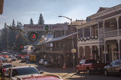 A festive street scene with a "Parade of Lights" banner, decorated buildings, and parked cars in a small town.