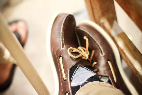 A close-up of brown shoes with laces, paired with patterned socks, resting on a floor near a chair.